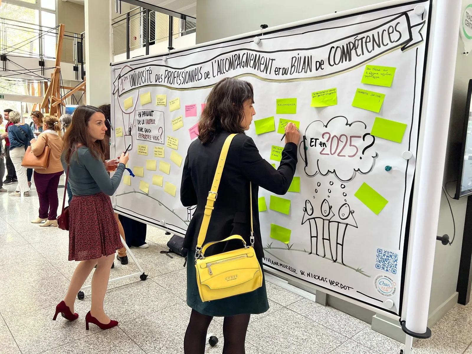 Deux participantes complètent une fresque murale : l’une écrit sur un post-it collé au panneau, l’autre tient un feutre