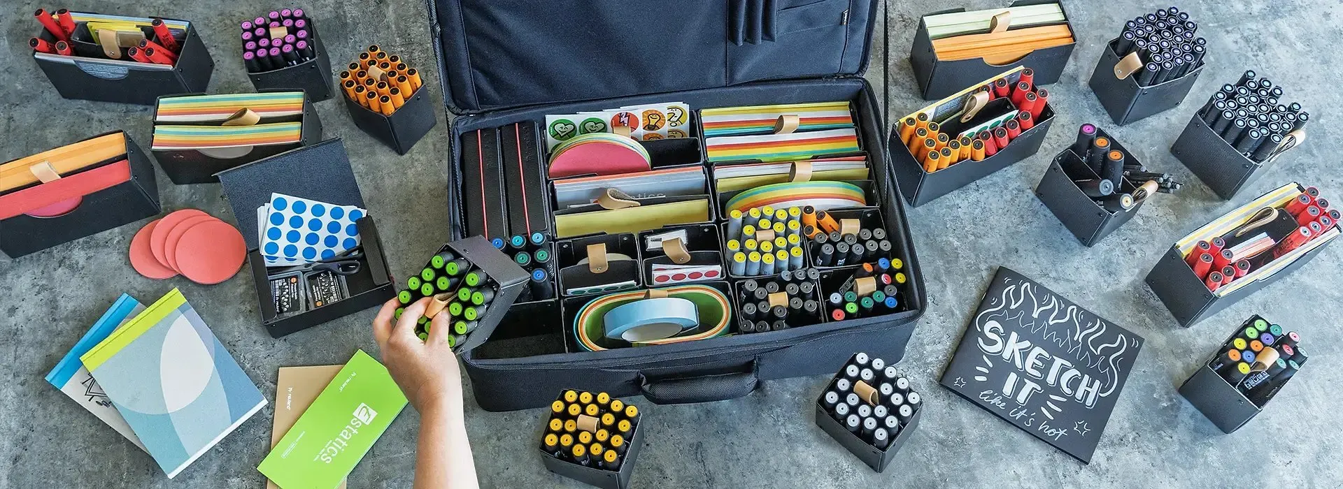 La valise Neuland ouverte sur un sol de béton, entourée de boîtes de marqueurs triées par couleur, de papiers, de gommettes et d'un carnet « Sketch it like it's hot »